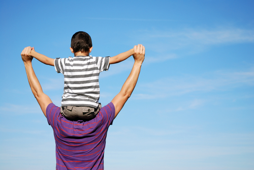 A child is sitting on the fathers shoulders who is carrying him around outdoors in the blue sky